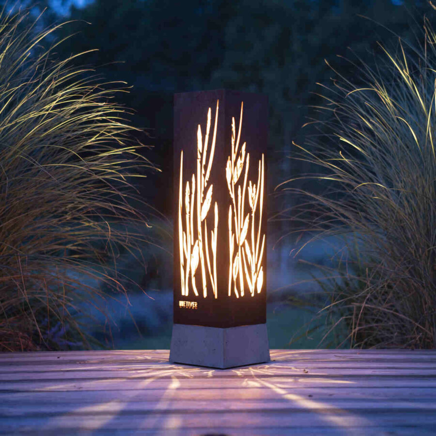 Lampa ze stali corten rozświetlająca drewniany taras otoczony trawami.